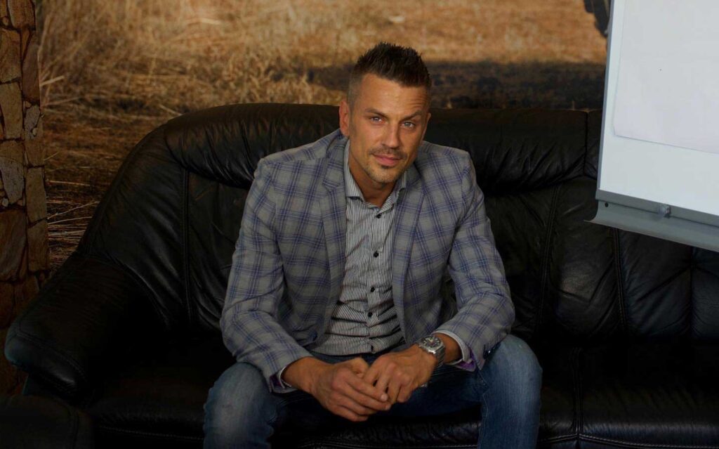 Mann in kariertem Sakko lächelnd auf Ledersofa, mit kurzem Haar und Uhr, vor rustikalem Hintergrund.