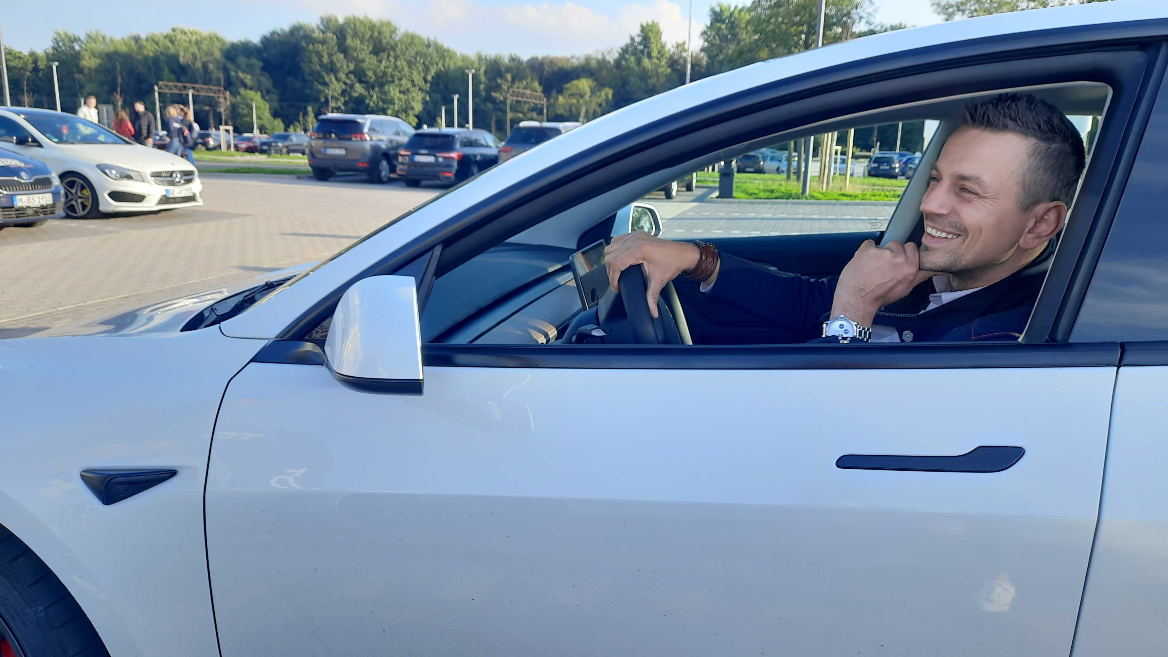 Mann im Auto lächelt; weißes Fahrzeug, sonniger Parkplatz im Hintergrund.