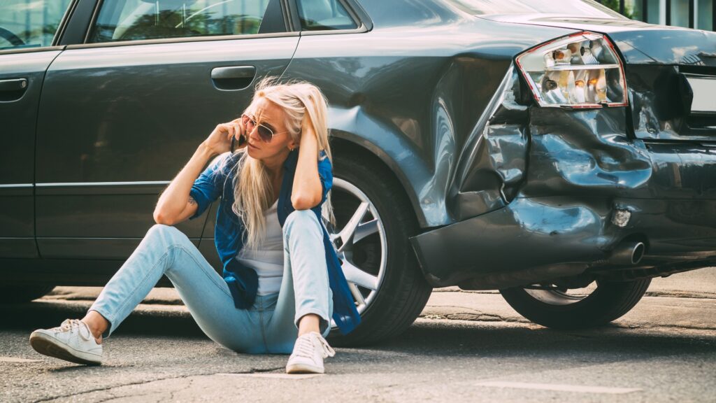 Verzweifelte Frau sitzt neben ihrem Unfallwagen und telefoniert