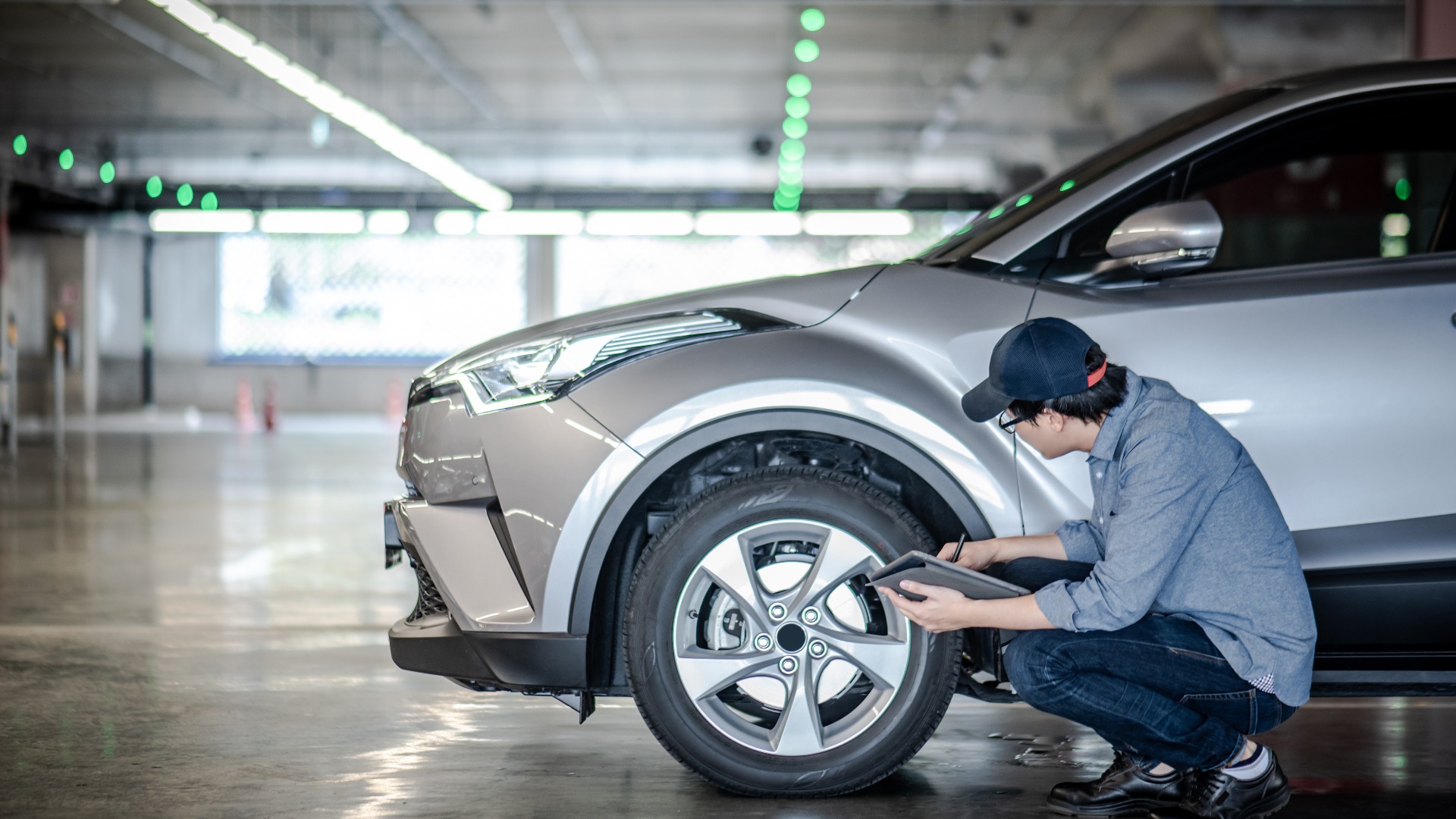 Ein Mechaniker prüft einen silbernen Wagen. Er hockt vor dem linken Vorderrad mit einer Checkliste.